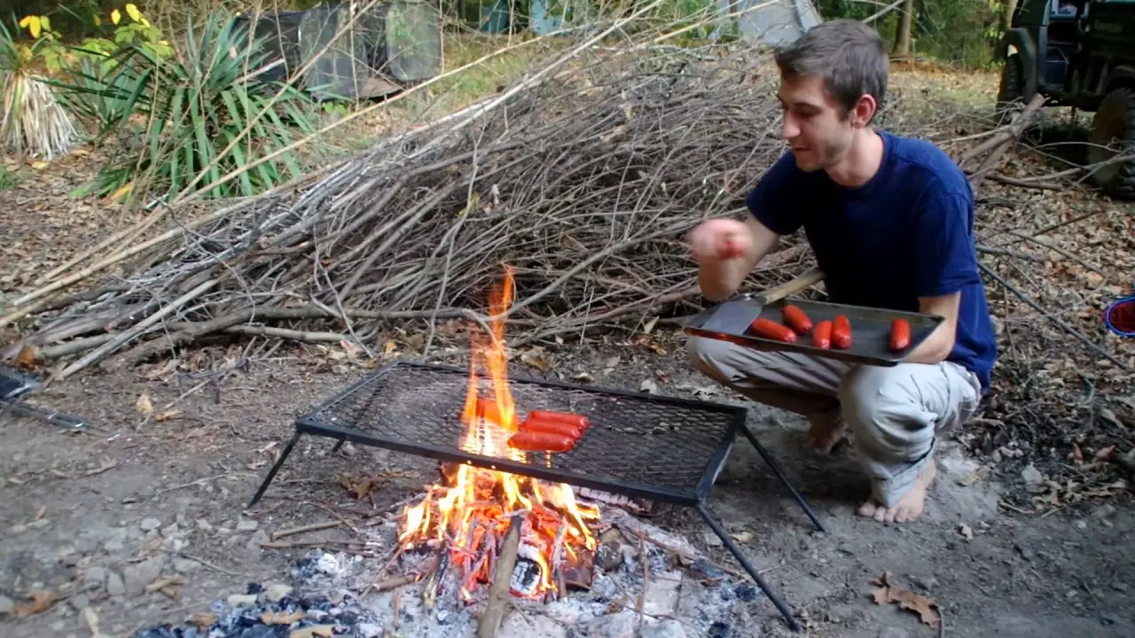 man leaning over Texsport campfire grill cooking hotdogs